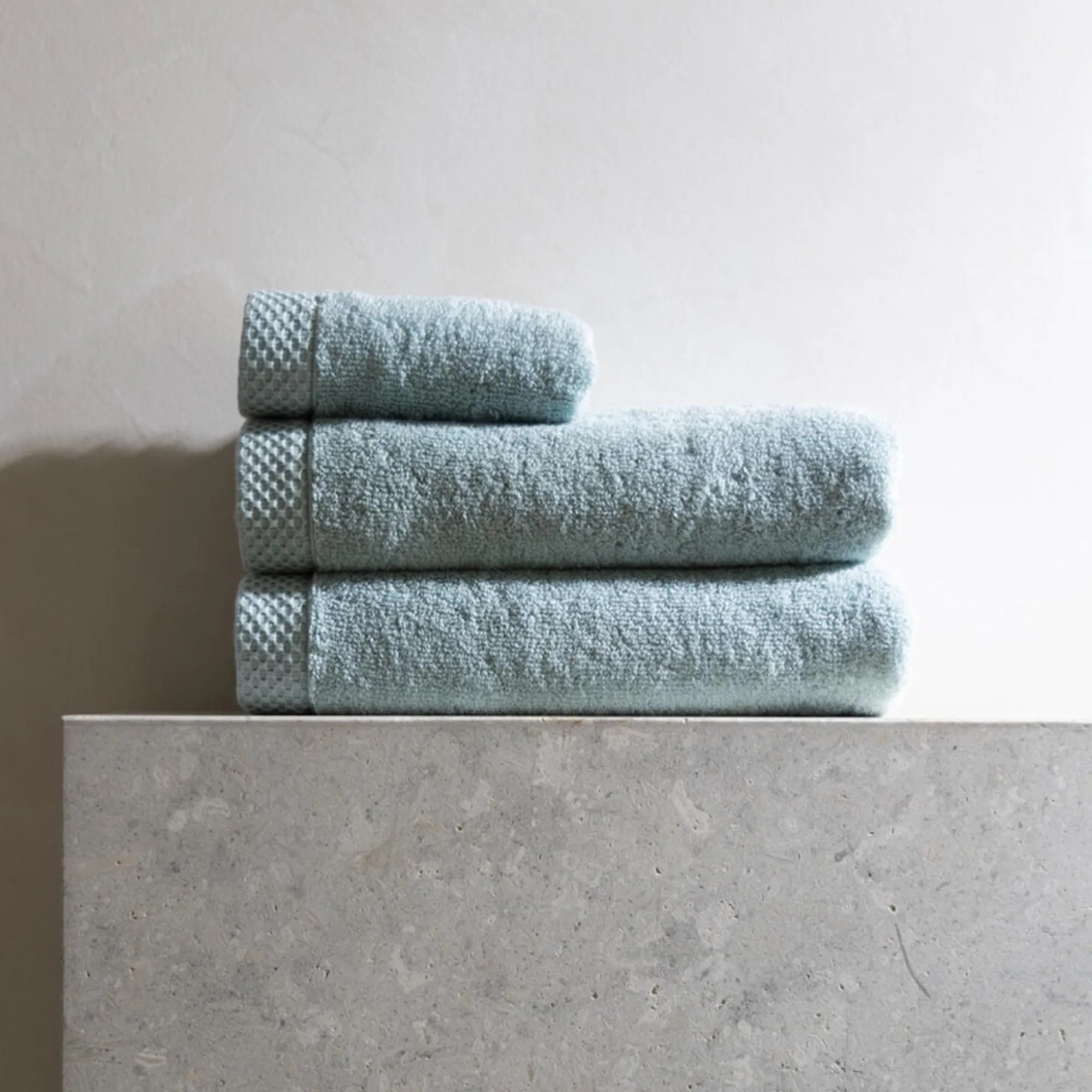 Drie op elkaar gevouwen luxe Florence-handdoeken in hemelsblauw, twee grote en één kleinere, liggend op een brede plank met betonlook