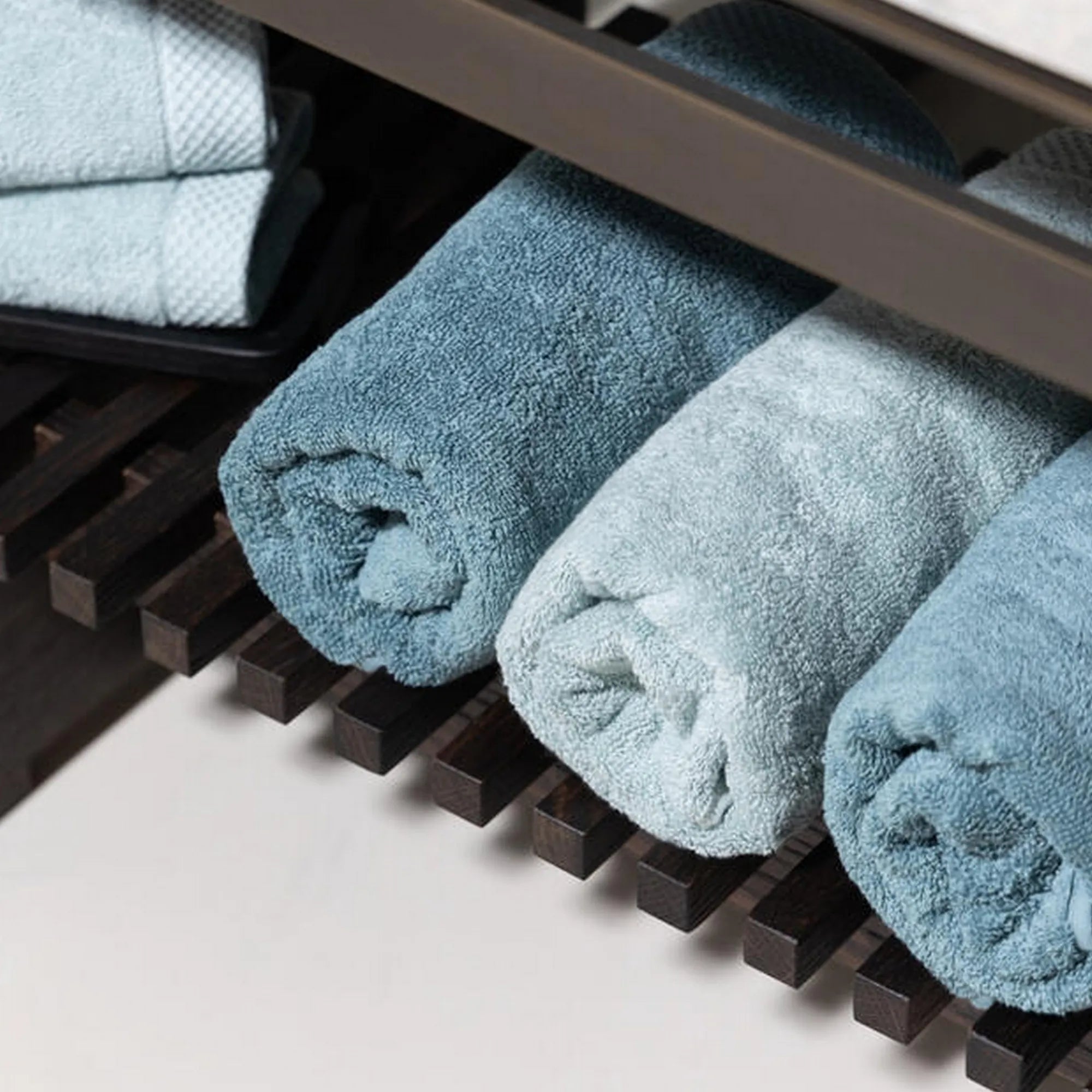 Drie opgerolde Florence-handdoeken in mineraalblauw, naast elkaar geplaatst in een houten badkamerrek